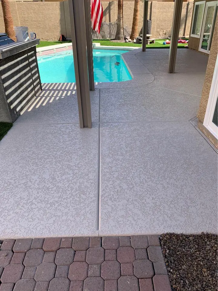 View of a backyard patio in San Bernardino featuring a swimming pool with clear blue water. The patio, crafted by licensed concrete contractors, boasts a combination of textured concrete and brick pavers. A pergola provides shaded seating near the pool, with an American flag hanging in the background.