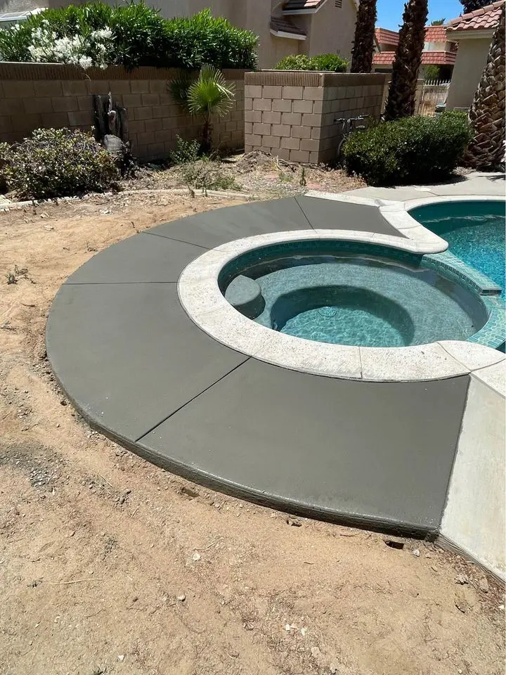 A freshly resurfaced concrete pool deck, expertly crafted by skilled pool remodeling contractors, surrounds a circular spa adjacent to a swimming pool filled with clear blue water. The yard features a tall concrete block wall, various plants, and a few trees in the background.