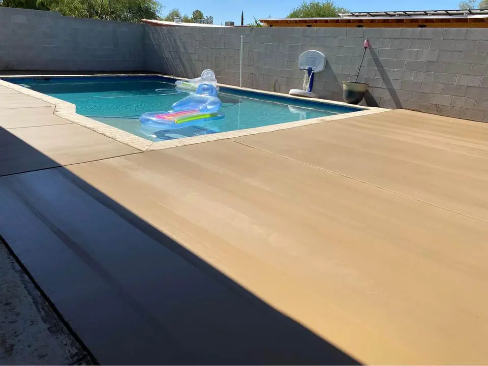 A backyard swimming pool in San Bernardino boasts crystal-clear water and a blue inflatable lounge float. Newly finished with a concrete pool deck, the area features a basketball hoop mounted on the far side of the gray cinder block wall, surrounded by trees visible in the background.