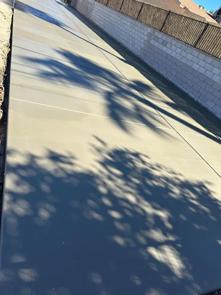 A freshly paved concrete path with shadows of tree branches cast on it. On one side is a cinder block wall topped with a wooden fence, and on the other side is a sloping dirt embankment. The scene, crafted by a licensed concrete contractor, is brightly lit by the sun.