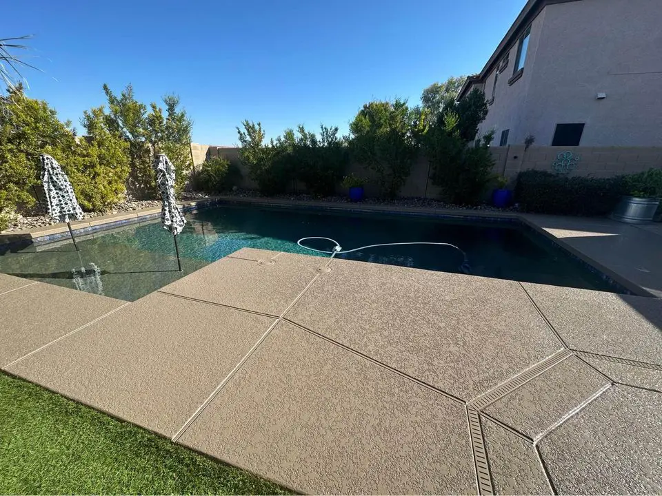 A backyard scene with a swimming pool surrounded by a concrete deck crafted by top-notch concrete pool deck contractors and well-maintained greenery. Two pool umbrellas are positioned near the shallow end of the pool. A house with a beige exterior and windows is visible on the right side, beneath a clear blue sky overhead.