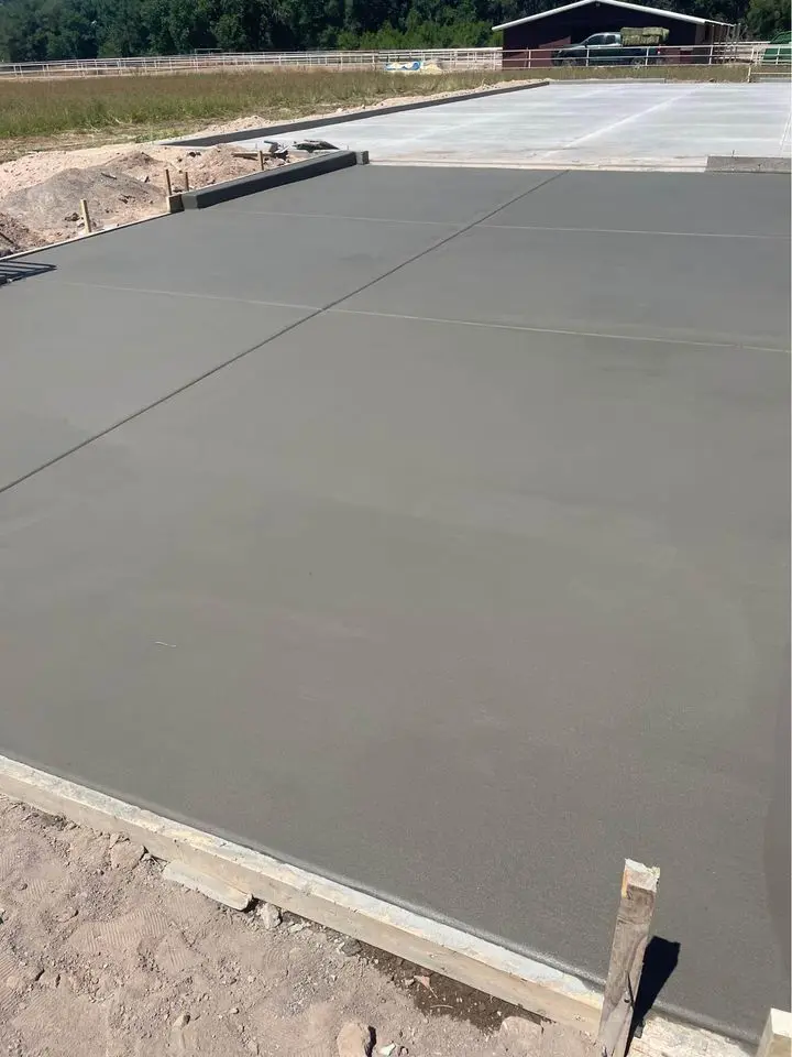 A freshly poured and smoothed concrete slab in an outdoor construction site, expertly prepared by skilled pad contractors. The surface is flat and evenly finished with visible joint lines. In the background, another concrete slab, a dirt area, and a fenced area with a building can be seen.