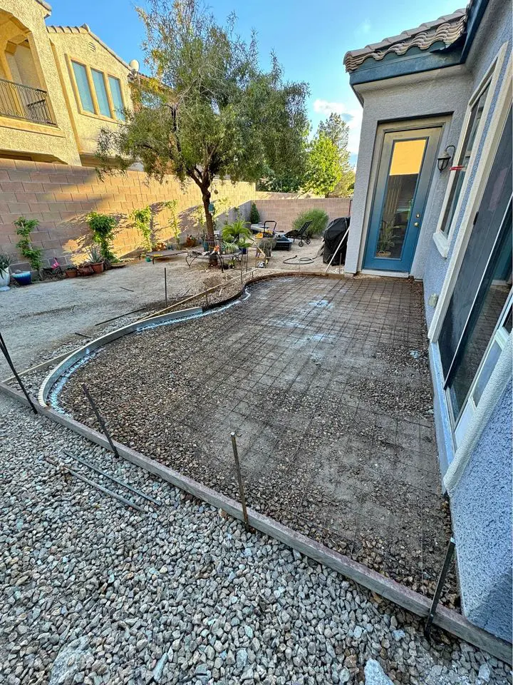 A backyard under renovation features a partially constructed concrete patio next to the house. The ground is covered with gravel, and metal stakes outline a future garden bed or pathway. Nearby, potted plants sit by a wooden fence, with neighboring houses in San Bernardino visible in the background.