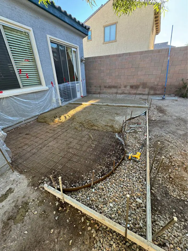 A backyard construction site with a partially completed patio in San Bernardino. There's a grid of rebar on the ground, surrounded by wooden forms and gravel. Plastic sheeting covers windows and edges of the house. A power tool from Berdoo Concrete rests near the rebar, reflecting skilled concrete patio contractors at work.