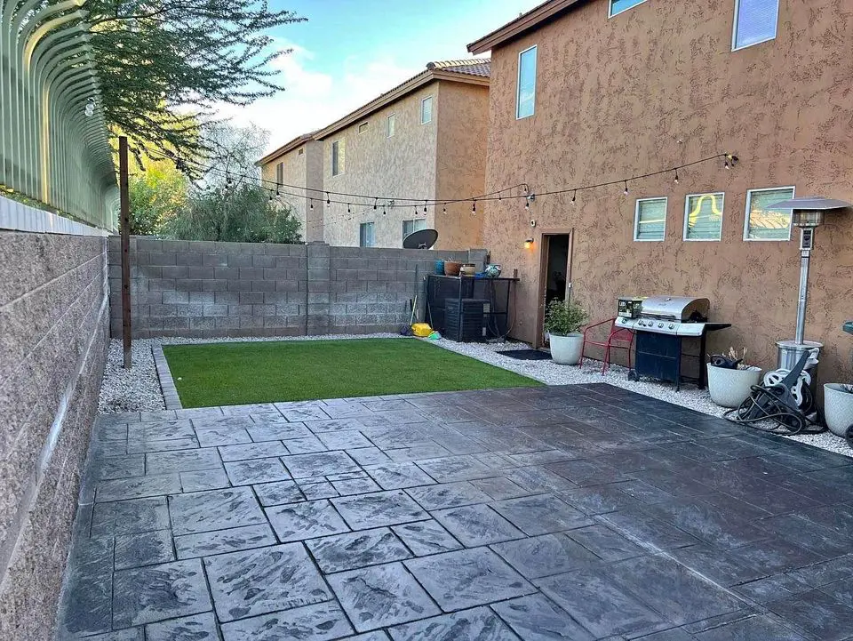 A backyard with a large paved patio area, crafted by expert concrete patio contractors, adjacent to a small grass lawn. The space is enclosed by a stone wall and features string lights, planters, a barbecue grill, and a patio heater. Two modern houses with stucco finish are visible in the background.