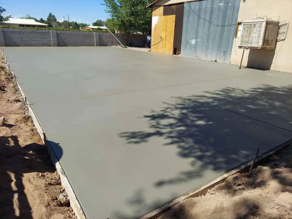 A freshly poured concrete slab in a yard, expertly done by San Bernardino's premier concrete contractors. Surrounded by a dirt area, it features a gray metal building with a wooden door and an air conditioning unit in the background. A cinder block wall encloses the area with trees and houses visible beyond it.