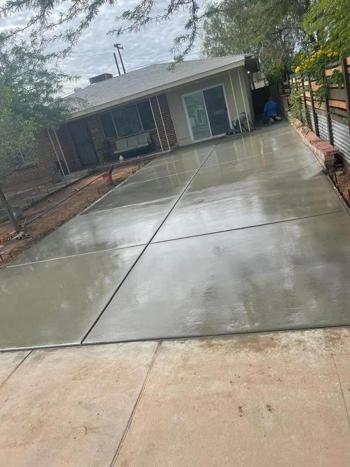 A freshly poured and smoothed concrete driveway in front of a brick house with large windows and a sliding door at the back. A concrete contractor from San Bernardino is nearby, with construction materials around. Trees and fencing line the sides of the driveway, giving it a polished look.