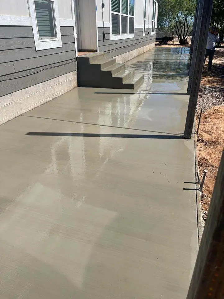 A freshly poured concrete driveway and sidewalk area outside a gray and white house with a set of steps leading up to an elevated entrance. The wet concrete surface is smooth, reflecting sunlight. Nearby, there is a tree and some construction materials, indicative of expert work by the local concrete contractor in San Bernardino, CA.