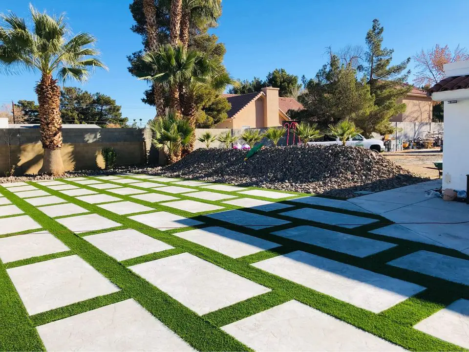 A backyard scene in San Bernardino, CA, featuring a modern patio design with alternating concrete tiles and green artificial grass. The area is bordered by palm trees and desert plants. A large pile of rocks and a white car are in the background against a sunny blue sky.