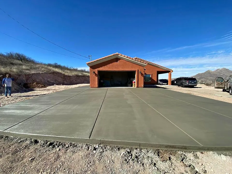 A newly poured concrete driveway by a licensed concrete contractor leads up to a brick house with two open garage doors. The sky is clear and blue, and there are hills in the background. Two people and a few vehicles are visible near the garage, enjoying their home in beautiful Loma Linda.