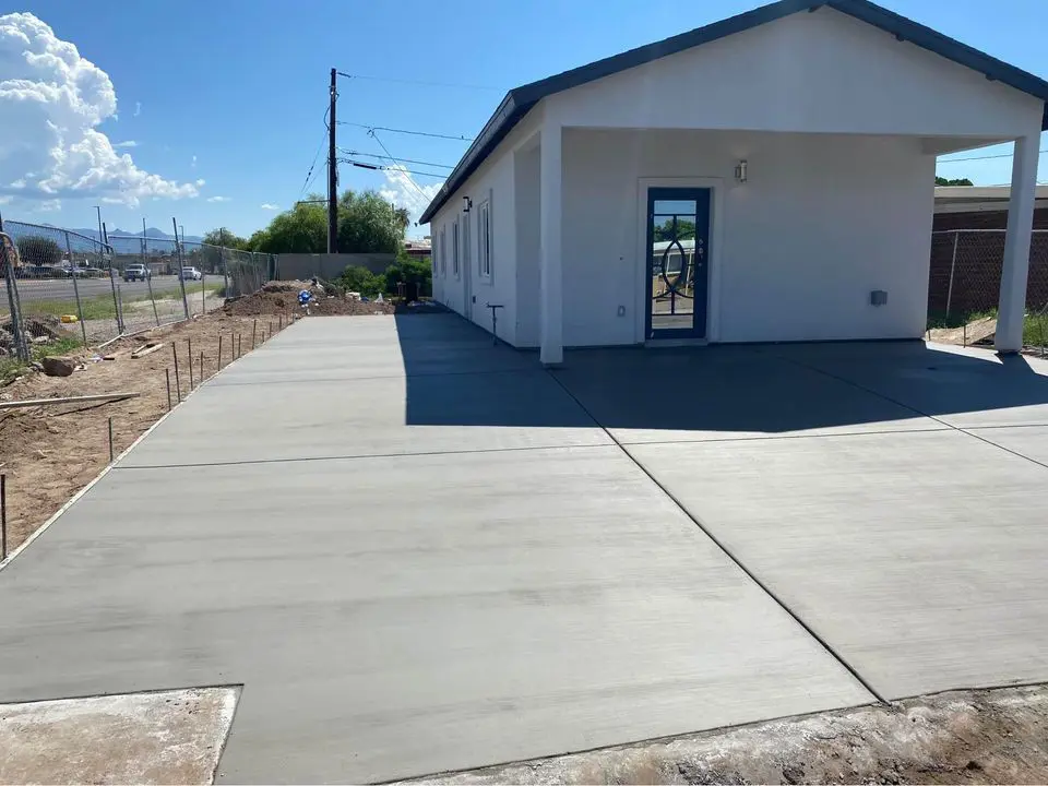 A small white house with a blue door stands beside a wide, freshly poured concrete driveway, thanks to a skilled concrete contractor in San Bernardino CA. The area around the house is mostly barren, with some construction materials and fencing visible. Trees are visible in the background under a clear blue sky.