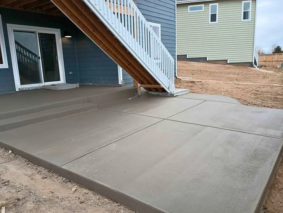 An outdoor concrete patio with fresh, smooth concrete extends from a house with blue siding and a set of stairs, reflecting the craftsmanship of Berdoo Concrete. The patio is still curing, with no visible furniture or decorations. Another house is visible in the background across a partially barren yard in San Bernardino.