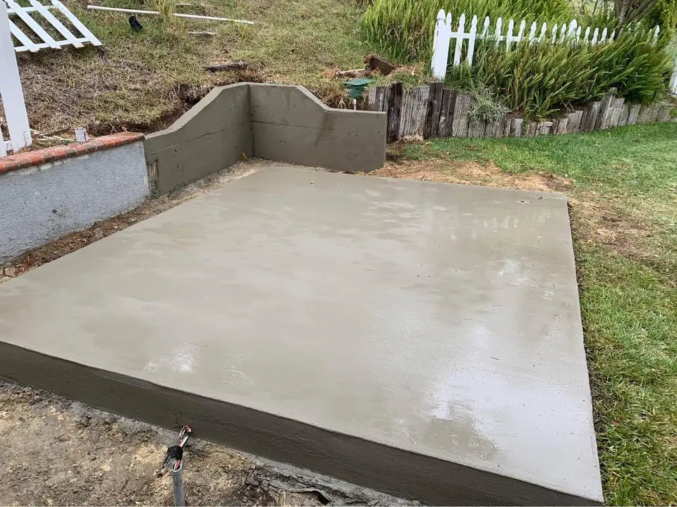 A small concrete slab with smooth edges has been freshly poured in a backyard by expert concrete slab contractors. It is adjacent to a short retaining wall and white picket fence. The surrounding area, typical of San Bernardino, includes patches of grass and dirt.