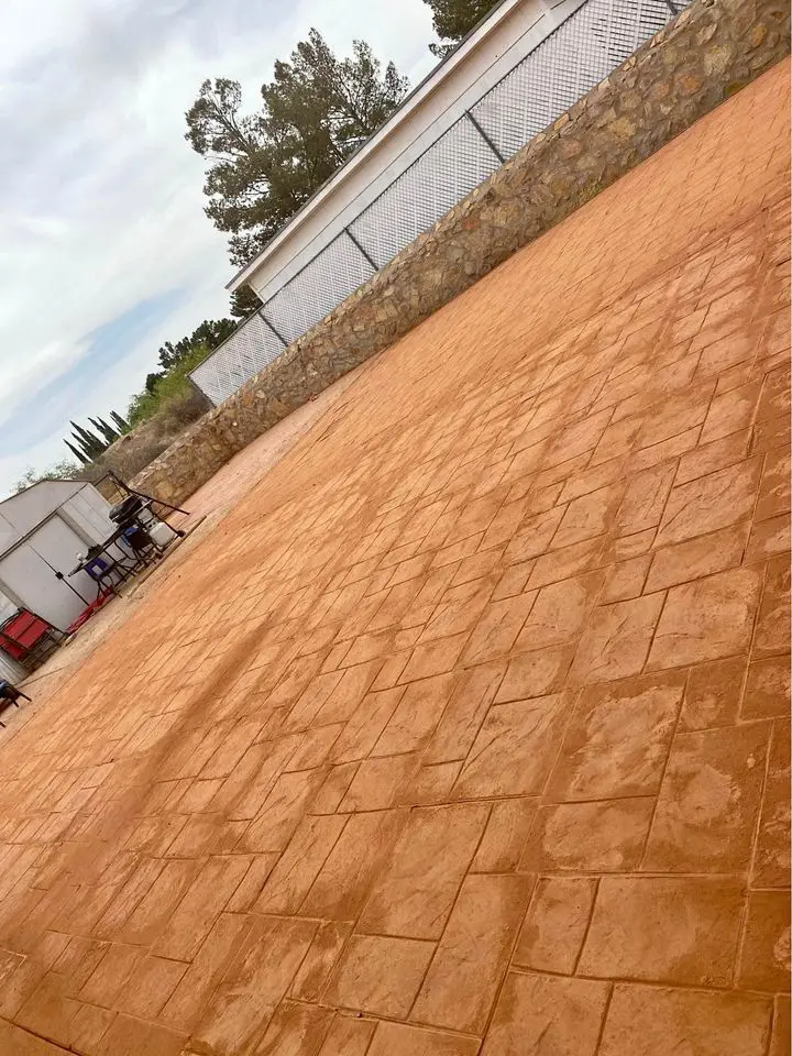 A spacious outdoor patio area is paved with reddish-brown tiles arranged in a geometric pattern. In the background, a stone wall with a chain-link fence on top borders the space, while trees and a cloudy sky are visible. Several pieces of equipment from our concrete contractor in San Bernardino, CA are off to the left side.