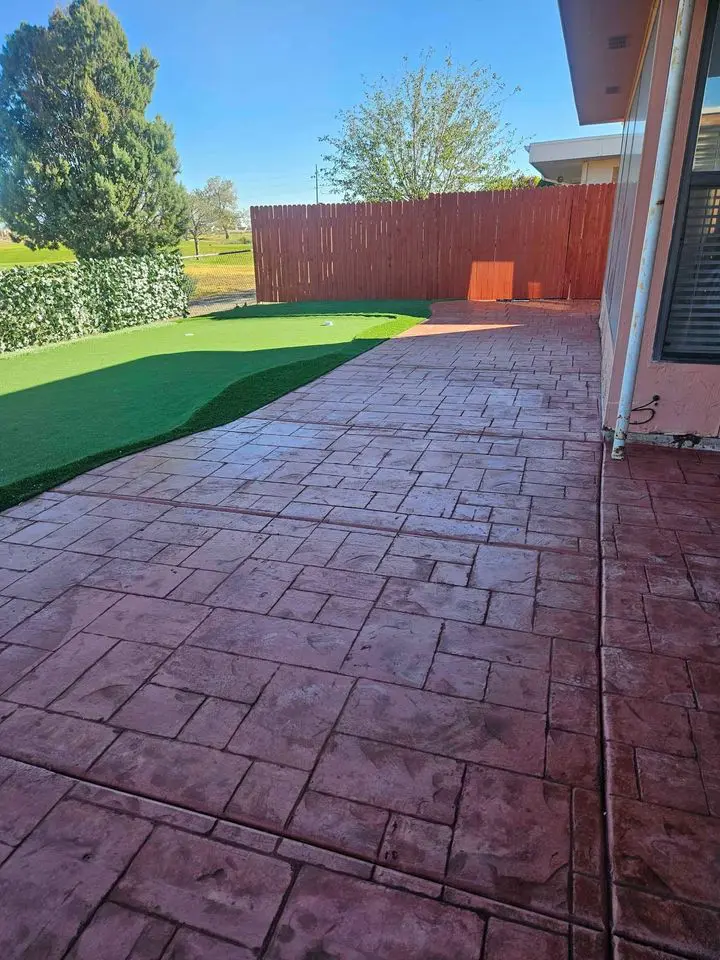 A backyard with a spacious, stamped concrete patio extending from the house. Adjacent to the patio is a section of artificial grass, bordered by a red wooden fence and lush greenery. Trees and a clear blue sky are visible in the background, beautifully crafted by top San Bernardino concrete patio contractors.