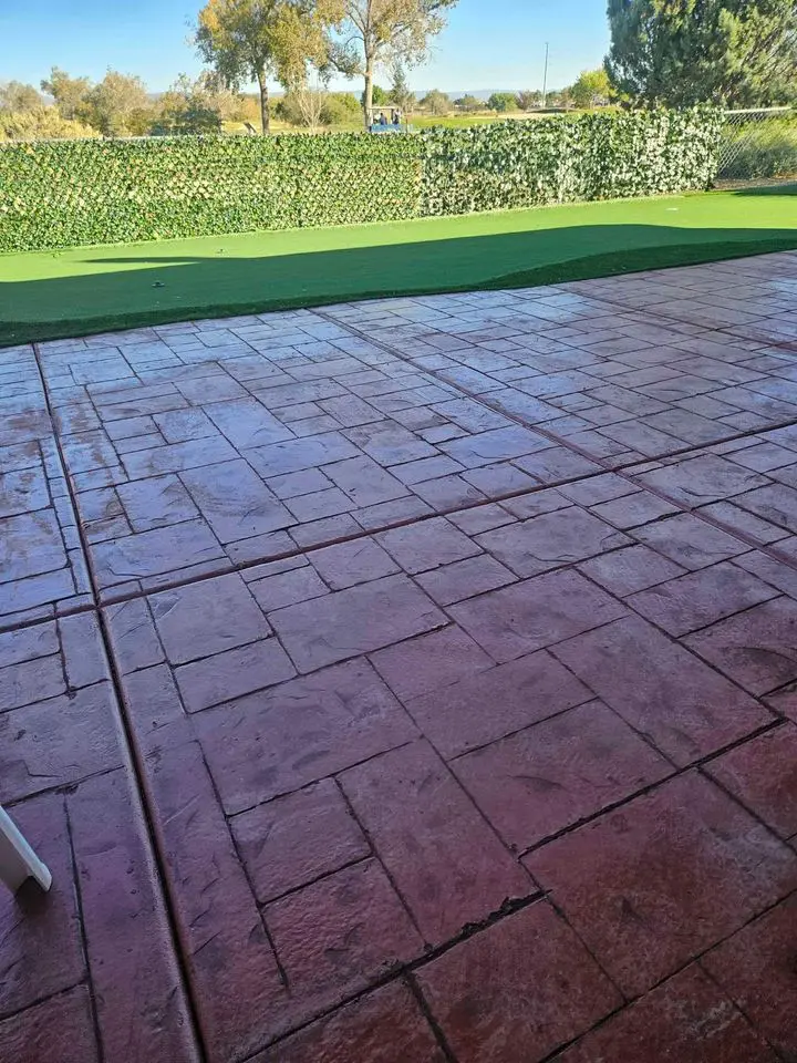 A large red brick patio, installed by Berdoo Concrete, glistens wet under the light, possibly from recent rain. Beyond the patio lies a stretch of green artificial turf, ending at a short hedge with lush foliage. Trees and a blue sky can be seen in the background of this Loma Linda residence.