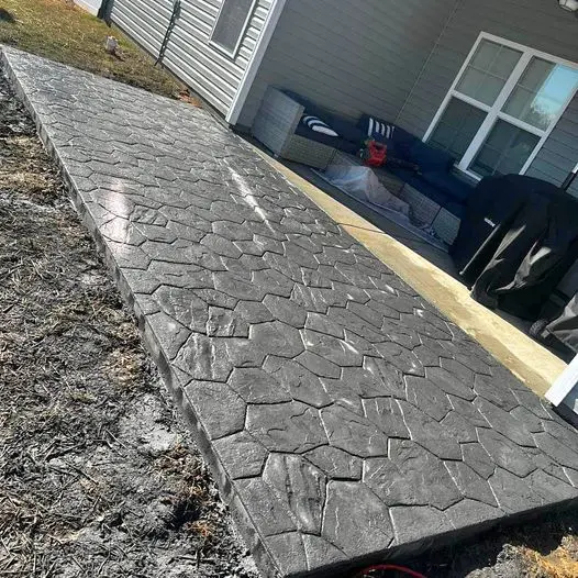 A newly installed concrete pathway with a hexagonal stone pattern by Berdoo Concrete leads to a covered patio area. The patio has white framed windows, outdoor furniture, and a grill. The surrounding ground appears unfinished, with some grass and dirt visible in Loma Linda.