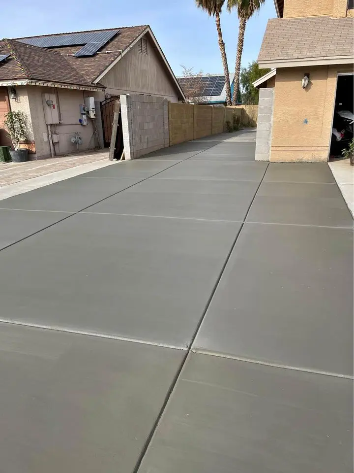 A long, freshly paved concrete driveway by Berdoo Concrete extends alongside a tan house with a garage. The driveway, neatly divided into sections by control joints, appears smooth and clean. In the background, you can see neighboring houses and palm trees typical of Loma Linda.