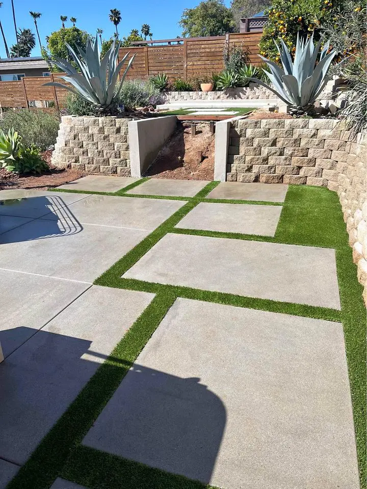 A modern outdoor patio with large, square concrete pavers bordered by green artificial grass, crafted by licensed concrete contractors from San Bernardino. Two large agave plants are placed symmetrically at the top of the stone steps, and a built-in fire pit is visible in the background. The sky is clear and sunny.