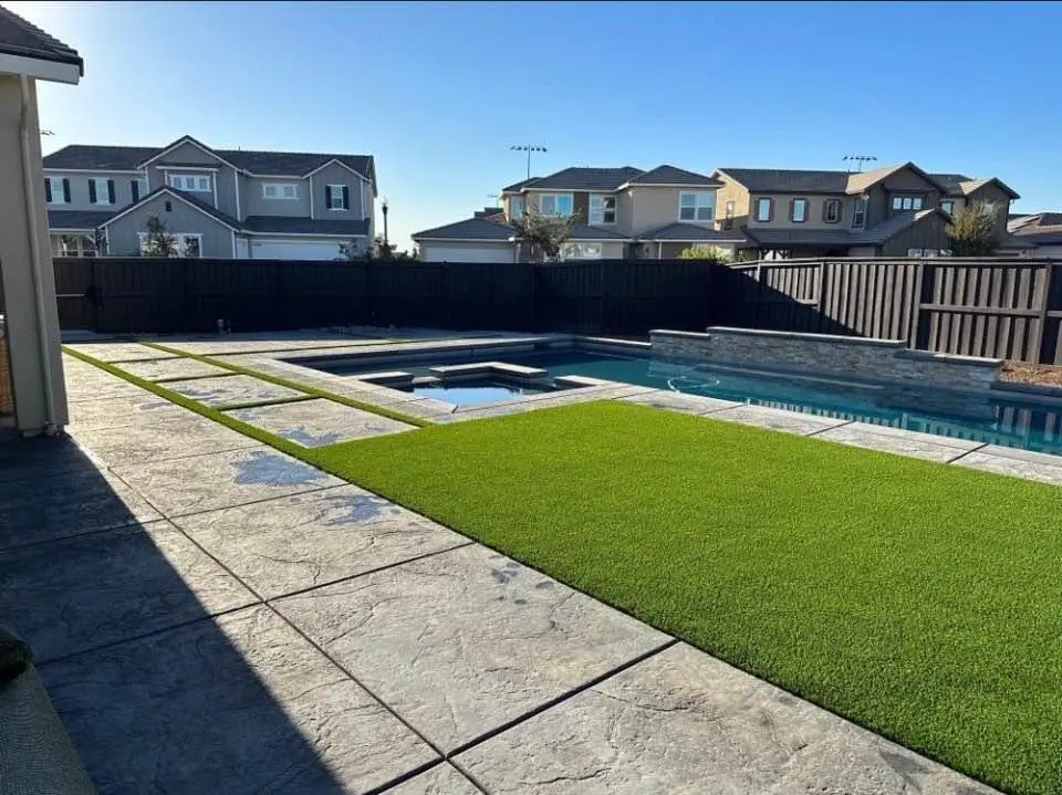 A modern backyard with a swimming pool and attached hot tub, both surrounded by green artificial turf. The area is enclosed by a dark wooden fence, crafted by reliable concrete services. Multiple two-story houses are visible in the background under a clear blue sky.