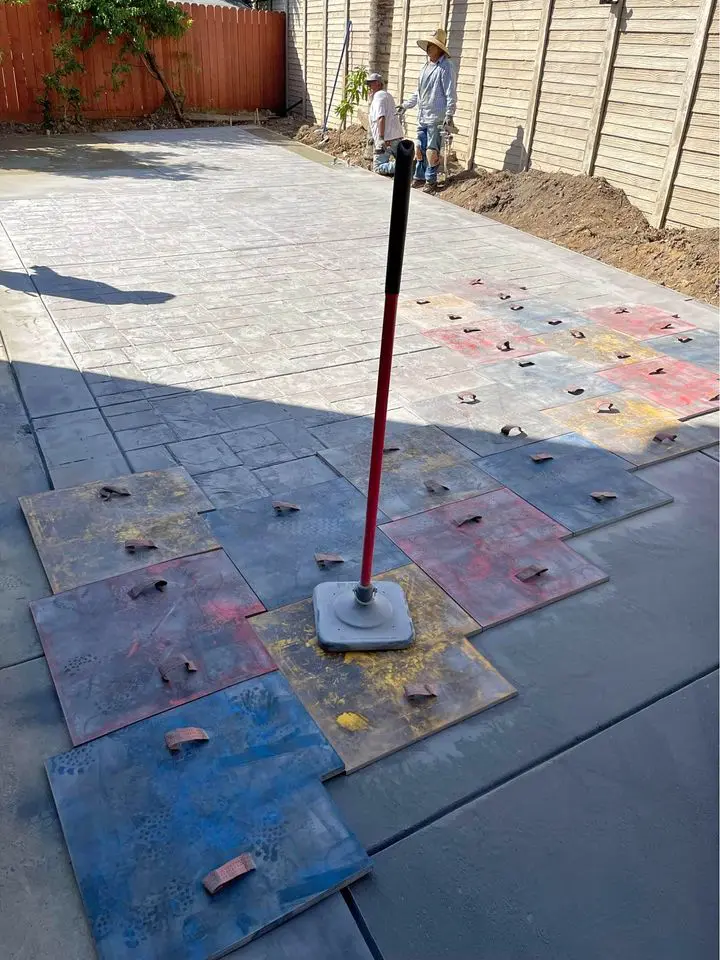 Two licensed concrete contractors at a San Bernardino construction site stand near a newly laid concrete surface. Mats with handles, used to imprint patterns into the concrete, are scattered on the ground. A tall tool with a handle is in the center of the image, likely used for imprinting.