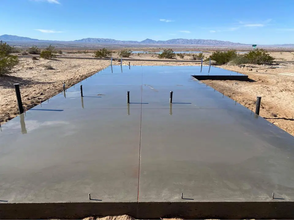 A newly poured concrete foundation slab sits in a desert landscape near San Bernardino. The smooth surface of the concrete reflects the blue sky. Small shrubs are scattered across the surrounding dry land, and distant mountains are visible on the horizon, showcasing expert work by licensed concrete contractors.