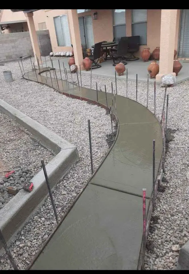 A curved concrete pathway is under construction in San Bernardino, framed by wooden forms and supported by metal stakes. The path, overseen by licensed concrete contractors, is surrounded by gravel and leads to a patio area with outdoor furniture. Several large pots are placed near the patio.