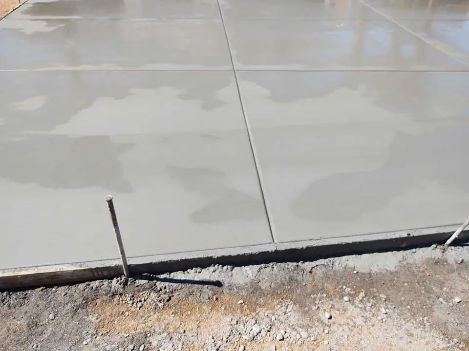 A freshly poured concrete slab, still wet and smooth, with square sections visible due to expansion joints. The surrounding area shows gravel and dirt, and a metal rod sticking out from the ground near the edge of the slab. Licensed concrete contractors in San Bernardino CA ensure top-quality concrete services like this.