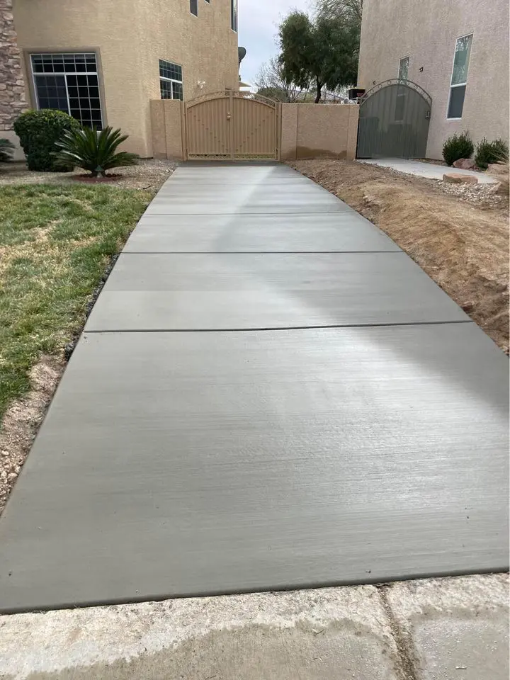 Freshly poured and smoothed concrete pathway leading between two residential buildings towards a closed double gate. The pathway is bordered by patches of grass and bare soil. The surrounding beige and light grey buildings showcase the expertise of licensed concrete contractors in San Bernardino.