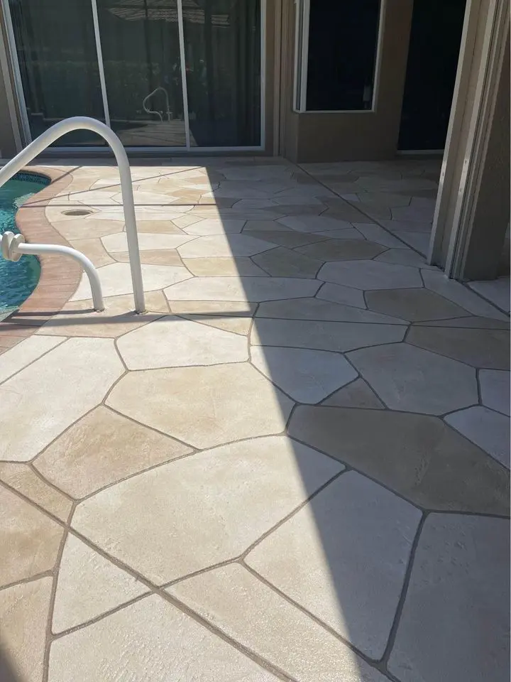 A sunlit poolside patio featuring large, irregularly-shaped stone tiles installed by licensed concrete contractors. A metal handrail curves over the pool's edge, casting shadows on the light tan and cream-colored tiles. Glass doors and windows are in the background, reflecting the outdoor area.