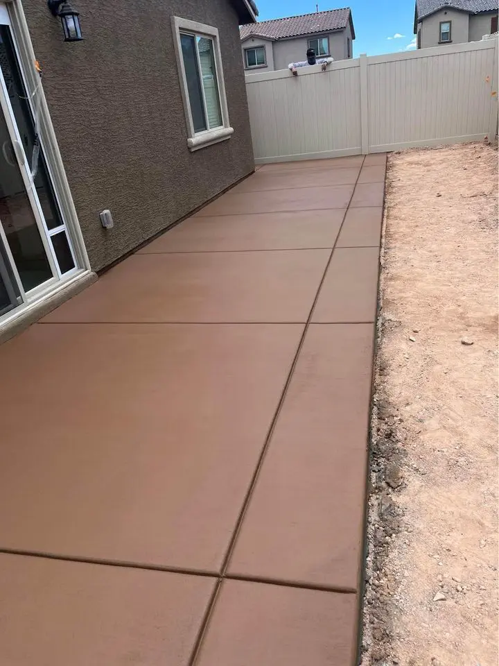 A freshly poured and smoothed concrete patio beside a house, bordered by dirt on one side and a white privacy fence on the other, crafted by licensed concrete contractors from Berdoo Concrete. The patio lies parallel to the house, with a sliding glass door visible on the left side of the image.