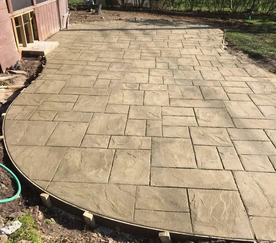 A newly constructed stamped concrete patio with a stone pattern curves around a brown wooden building in San Bernardino. The surrounding area is under construction, showing exposed soil and patches of green grass at the edges. A garden hose lies in the bottom-left corner, ready for use by licensed concrete contractors.