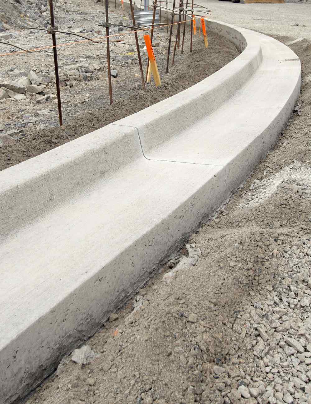 A fresh, curved concrete curb, crafted by licensed concrete contractors, is shown in daylight at a San Bernardino construction site. The area around the curb is rough and unpaved, with dirt and scattered rubble indicating ongoing work. In the background, temporary scaffolding and construction markers are visible.